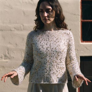 May include: A long-sleeved, cream-colored fuzzy knit sweater featuring a round neckline and scattered gold sequins throughout the fabric. The sweater has a relaxed fit with slightly flared sleeves and a soft, textured appearance.