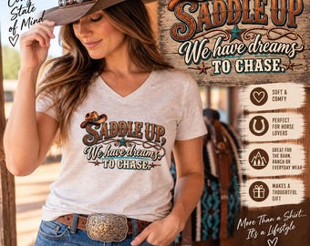 Könnte beinhalten: Ein hellgrau meliertes V-Ausschnitt-T-Shirt mit einem Grafikdruck eines braunen Cowboyhuts, eines Lassos und dem Text "SADDLE UP, We have dreams TO CHASE." Das T-Shirt wird mit einer blauen Jeans und einer großen, verzierten Metallschnalle kombiniert.