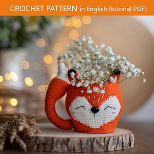May include: A crochet pattern tutorial PDF titled CROCHET PATTERN in English. The image features a fox-shaped crochet mug cozy in orange and white, with a fox tail handle, black eyes, and small ears, filled with white baby's breath flowers.