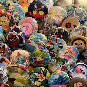 Op de afbeelding: Een verzameling ronde buttons met diverse kleurrijke stripfiguren, waaronder wezens met opvallende kenmerken zoals puntige oren, grote ogen en uiteenlopende patronen, gerangschikt in een dichte, overlappende groep.