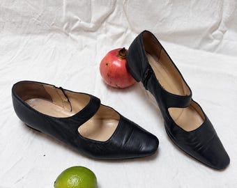Puede incluir: Un par de zapatos Mary Jane de cuero negro con tacón bajo, punta cuadrada y una tira sobre el empeine. Los zapatos están colocados junto a una granada entera y una lima verde, resaltando la textura suave del cuero negro.