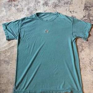 May include: A short-sleeved crew neck t-shirt in a muted teal green color. The shirt features a small, centered embroidered logo on the chest that displays the letters "OF" in a colorful, rainbow-like gradient pattern against the solid fabric.