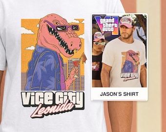 May include: A white t-shirt featuring a graphic of a pink alligator wearing purple sunglasses and a blue palm-print shirt, holding a cocktail. Below the graphic, the text reads Vice City Leonida. A small inset shows a person wearing the same shirt.