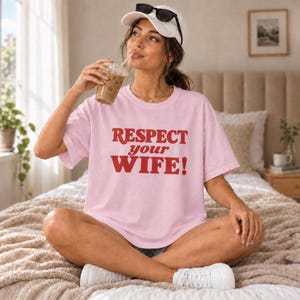 Könnte beinhalten: Ein hellrosa T-Shirt mit kurzen Ärmeln und dem roten Schriftzug "RESPECT your WIFE!" auf der Vorderseite. Das Oberteil hat einen Rundhalsausschnitt und eine lockere Passform, gefertigt aus einfarbigem Stoff mit kräftigen Großbuchstaben.