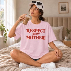 Puede incluir: Una camiseta de manga corta de color rosa claro con el texto "RESPECT your MOTHER!" impreso en letras rojas llamativas en el pecho. La prenda cuenta con un cuello redondo y un corte holgado para un estilo casual y cotidiano.
