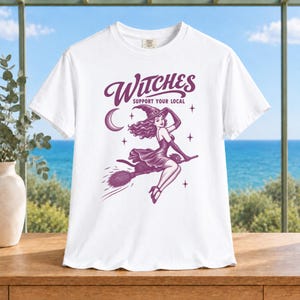 May include: A white short-sleeved t-shirt featuring a purple graphic print of a person wearing a witch hat and dress, riding a broomstick. The text above the illustration reads "WITCHES SUPPORT YOUR LOCAL" with a crescent moon and stars surrounding the figure.