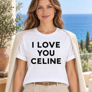 Può includere: Una t-shirt bianca a maniche corte con il testo nero I LOVE YOU CELINE stampato sul davanti. La maglietta è indossata con un blazer beige chiaro appoggiato sulle spalle e pantaloni beige con una sottile cintura marrone con fibbia dorata.