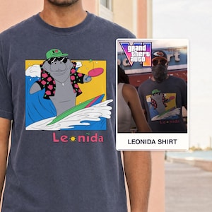 Op de afbeelding: Een donkerblauw t-shirt met een afbeelding van een zeekoe die een groene pet, zonnebril en bloemetjeshemd draagt terwijl hij op een golf surft. De zeekoe houdt een roze peddel vast. Onder de afbeelding staat de tekst Leonida in roze letters.