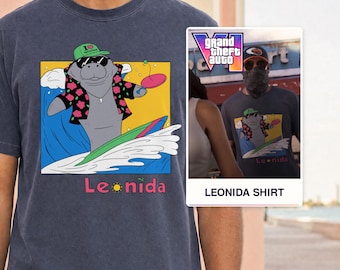 Puede incluir: Una camiseta azul oscuro con un gráfico de un manatí con gorra verde, gafas de sol y camisa de flores mientras surfea sobre una ola. El manatí sostiene una paleta rosa. Debajo del gráfico, el texto Leonida está impreso en letras rosas.