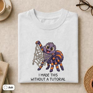 May include: A light gray t-shirt featuring a cartoon purple and orange spider holding a white web. Below the spider, the text reads: I MADE THIS WITHOUT A TUTORIAL. The design is centered on the chest of the shirt, which is laid flat on a neutral surface.