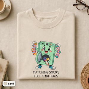 May include: A sand-colored t-shirt featuring a cartoon illustration of a green washing machine with a face, arms, and legs. The machine holds a striped sock and a polka-dot sock, with the text "MATCHING SOCKS FELT AMBITIOUS" printed below.