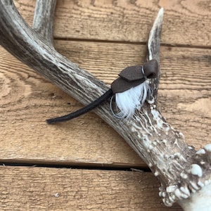 May include: A dark brown leather fly fishing lure featuring a small tuft of white feathers and a thin dark brown leather tail, positioned on a textured deer antler. The lure includes a curved metal hook visible beneath the white feathers.