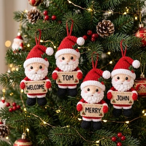 Op de afbeelding: Vier gehaakte kerstman-ornamenten hangend in een kerstboom. Elke kerstman draagt een rode muts en pak met zwarte laarzen en houdt een beige bordje vast met de teksten WELCOME, TOM, MERRY en JOHN in zwarte letters.