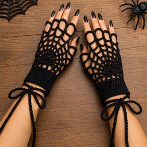 Puede incluir: Un par de guantes negros sin dedos con un patrón de telaraña de ganchillo. Los guantes tienen un puño sólido en la muñeca y cordones largos y finos de ganchillo que se envuelven alrededor de la muñeca y se aseguran con un lazo.
