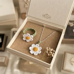 May include: A crochet daisy jewelry set featuring a ring and a necklace with a silver-tone chain, both with white petals and yellow centers, presented in a beige gift box with the text Gift Center Boutique printed on the inner lid.