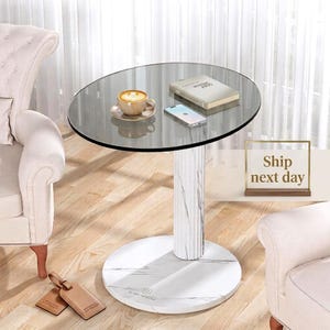 May include: A round side table with a glass top and a white fluted pedestal base featuring grey marble veining. The table holds a ceramic coffee cup on a saucer, a rectangular book, and a smartphone. The base sits on a circular marble platform.