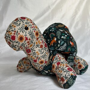 May include: A stuffed turtle toy featuring a head and limbs covered in a cream fabric with a red, orange, and green floral pattern. The shell is made of a dark green fabric decorated with a print of orange foxes, rainbows, and mushrooms.