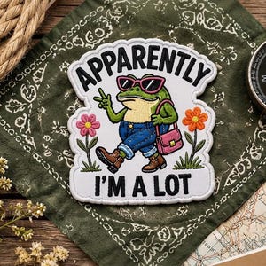May include: A white embroidered patch featuring a green frog wearing pink sunglasses, blue jeans, and brown boots, carrying a pink shoulder bag. The frog makes a peace sign. The text reads APPARENTLY I'M A LOT, with pink and orange flowers.