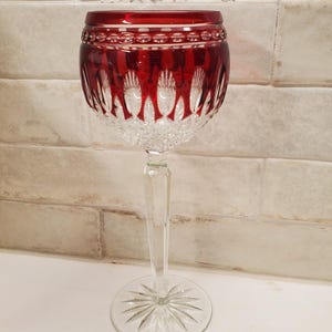 May include: A tall crystal wine glass featuring a deep red upper bowl with intricate cut glass patterns, including vertical arches and a diamond-cut base. The stem is clear and faceted, leading to a circular foot with a starburst design etched underneath.