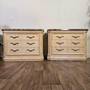 May include: A pair of matching three-drawer chests featuring a light beige wood grain finish, dark brown marble-look tops, and curved dark metal drawer pulls. Each chest has a rectangular frame with a wide base and three horizontal drawers.