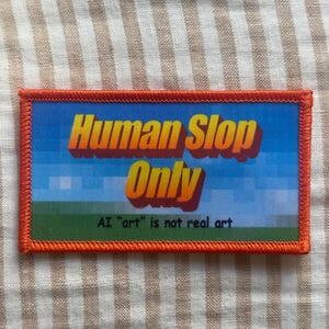 May include: A rectangular embroidered patch with an orange border. The center features a blue sky and green grass background with the yellow text "Human Slop Only" and the smaller black text "AI "art" is not real art" printed below.