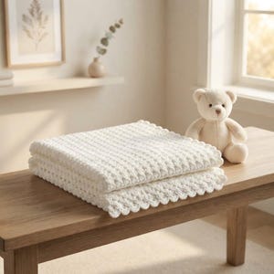 Peut inclure: Une couverture en crochet blanc pliée avec un motif texturé et un bord festonné, placée à côté d'un petit ours en peluche beige clair. La couverture présente un motif de points répétitif et est soigneusement empilée sur une surface en bois.