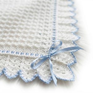 Può includere: Una coperta all'uncinetto bianca con un motivo a punti testurizzati e un bordo smerlato rifinito in azzurro. Un nastro di raso azzurro è intrecciato lungo il bordo e annodato in un fiocco all'angolo del pezzo tessile quadrato.