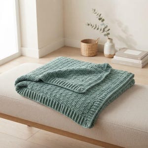 May include: A sage green knitted throw blanket with a textured, repeating square pattern and a ribbed border. The blanket is folded neatly, showcasing the soft, chunky yarn construction and the consistent, uniform stitch detail throughout the fabric.