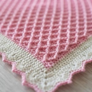 May include: A soft pink crochet blanket featuring a raised waffle stitch pattern throughout the center. The blanket is finished with a wide, cream-colored border that has a scalloped, decorative edge along the entire perimeter of the textile.
