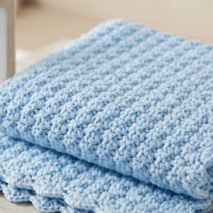 May include: A light blue crocheted blanket featuring a textured, repeating stitch pattern and a scalloped edge. The yarn is a soft, solid pastel blue color, and the blanket is folded into layers, showcasing the intricate, raised knit-like texture.