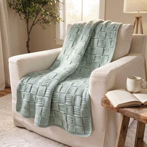 May include: A light sage green knitted throw blanket featuring a textured basketweave pattern. The blanket is draped over a cream-colored upholstered armchair, showcasing the thick yarn construction and the repeating square woven design.