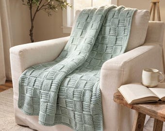 May include: A light sage green knitted throw blanket featuring a textured basketweave pattern. The blanket is draped over a cream-colored upholstered armchair, showcasing the thick yarn construction and the repeating square woven design.