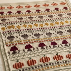 May include: A rectangular crochet blanket featuring horizontal rows of autumn-themed motifs including orange pumpkins, dark red mushrooms, brown acorns, and yellow leaves on a cream-colored background with decorative green and brown borders.