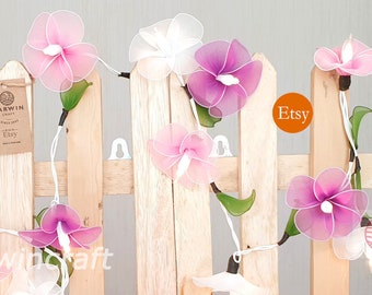 May include: String lights with pink and white fabric flowers, the lights are strung on a white wire and are hanging on a wooden fence. The text "Marwin Craft Etsy" is visible on the image.