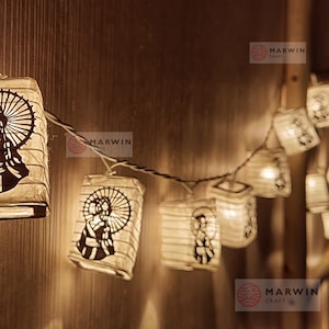 Puede incluir: Cadena de luces con faroles de papel que presentan la silueta de una persona sosteniendo un paraguas. Los faroles son blancos y emiten un brillo cálido. Las luces están enhebradas en un cable blanco. El texto "Etsy" y "Marwin Craft" es visible en la imagen.