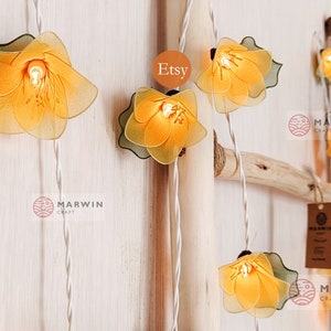 Puede incluir: Luces de cadena con pantallas de tela en forma de flor amarilla. Las luces están enhebradas en un cable blanco y cuelgan de una escalera de madera. El texto "Etsy" y "Marwin Craft" es visible en la imagen.