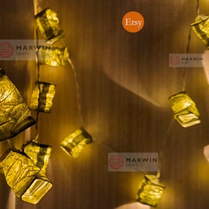 Puede incluir: Cadena de luces con pequeñas linternas de papel cuadradas de color dorado. Las linternas están encendidas y emiten un brillo cálido. Las luces están enhebradas en un cable blanco y cuelgan de una pared de madera. El texto "MARWIN CRAFT" es visible en la pared.