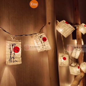 Op de afbeelding: Een string met witte papieren lampionnen met een rode cirkel en een bamboe ontwerp. De lampionnen zijn verlicht met warm witte LED-verlichting en zijn bevestigd aan een wit snoer. De lampionnen hangen aan een houten muur.