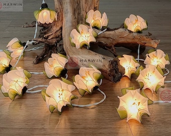 May include: A string of white flower lights with green leaves. The lights are lit and glowing. The lights are strung on a white wire. The lights are on a wooden surface. The lights are a decorative item for a home or party.