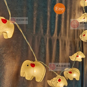 Könnte beinhalten: Eine Lichterkette mit weißen Elefanten-förmigen Lichtern mit roten Akzenten. Die Lichter sind an einem durchsichtigen Draht befestigt und leuchten. Die Lichter hängen an einer strukturierten Wand. Der Text "Etsy" und "MARWIN CRAFT" ist auf dem Bild zu sehen.