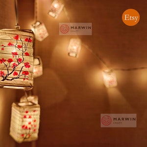 May include: String of white paper lanterns with pink cherry blossom designs, lit with warm white LED lights. The lanterns are hanging from a wire strung across a wall. The text "MARWIN CRAFT" is visible on the wall.