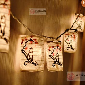 May include: String of illuminated paper lanterns. Each lantern is rectangular and features a black illustration of a cat on a branch with red flowers. The lanterns are strung together with a thin wire, creating a warm, inviting glow.