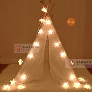 May include: A white teepee tent with a string of star-shaped lights wrapped around it. The lights are on and glowing. The teepee is on a white fluffy rug. The words "Marwin Craft" are visible on the left side of the image. The word "Etsy" is visible on the right side of the image.