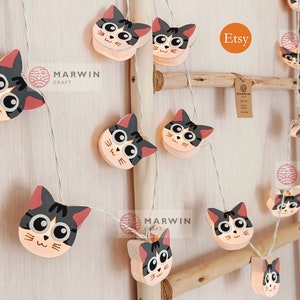May include: A string of white fairy lights with cat-shaped light covers. The lights are strung on a wooden ladder. The cat-shaped light covers are pink and white with black eyes and whiskers. The lights are labeled "Marwin Craft" and "Etsy".