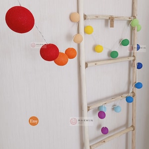 May include: A string of cotton ball lights in rainbow colours is draped over a wooden ladder. The lights are strung together with a clear wire. The ladder is made of natural wood and has four rungs. The lights are a popular decoration for homes and parties.