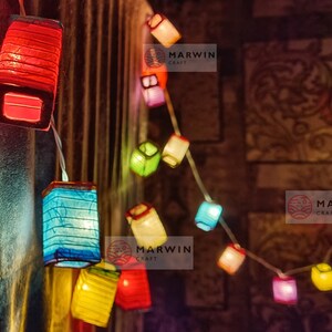 May include: A string of colorful paper lanterns in red, blue, yellow, and green. The lanterns are square-shaped and illuminated, strung on a white wire. The background is a blurred, patterned wall.