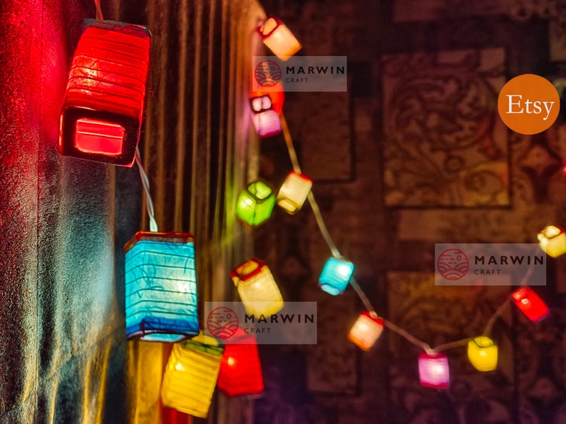 May include: A string of colorful paper lanterns in red, blue, yellow, and green. The lanterns are square-shaped and illuminated, strung on a white wire. The background is a blurred, patterned wall.