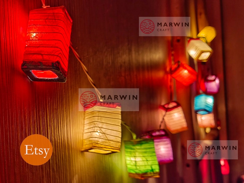 May include: A string of colorful paper lanterns with warm white lights. The lanterns are made of thin, translucent paper and are strung together with a clear wire. The lanterns are hanging on a wooden wall. The text "Etsy" and "Marwin Craft" is visible in the image.