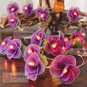 May include: Purple fabric flower string lights with warm white LED lights. The lights are strung on a clear wire and are perfect for adding a touch of whimsy to any space.