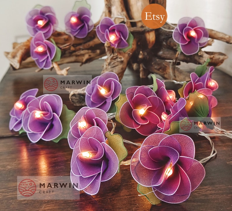 May include: Purple fabric flower string lights with warm white LED lights. The lights are strung on a clear wire and are perfect for adding a touch of whimsy to any space.